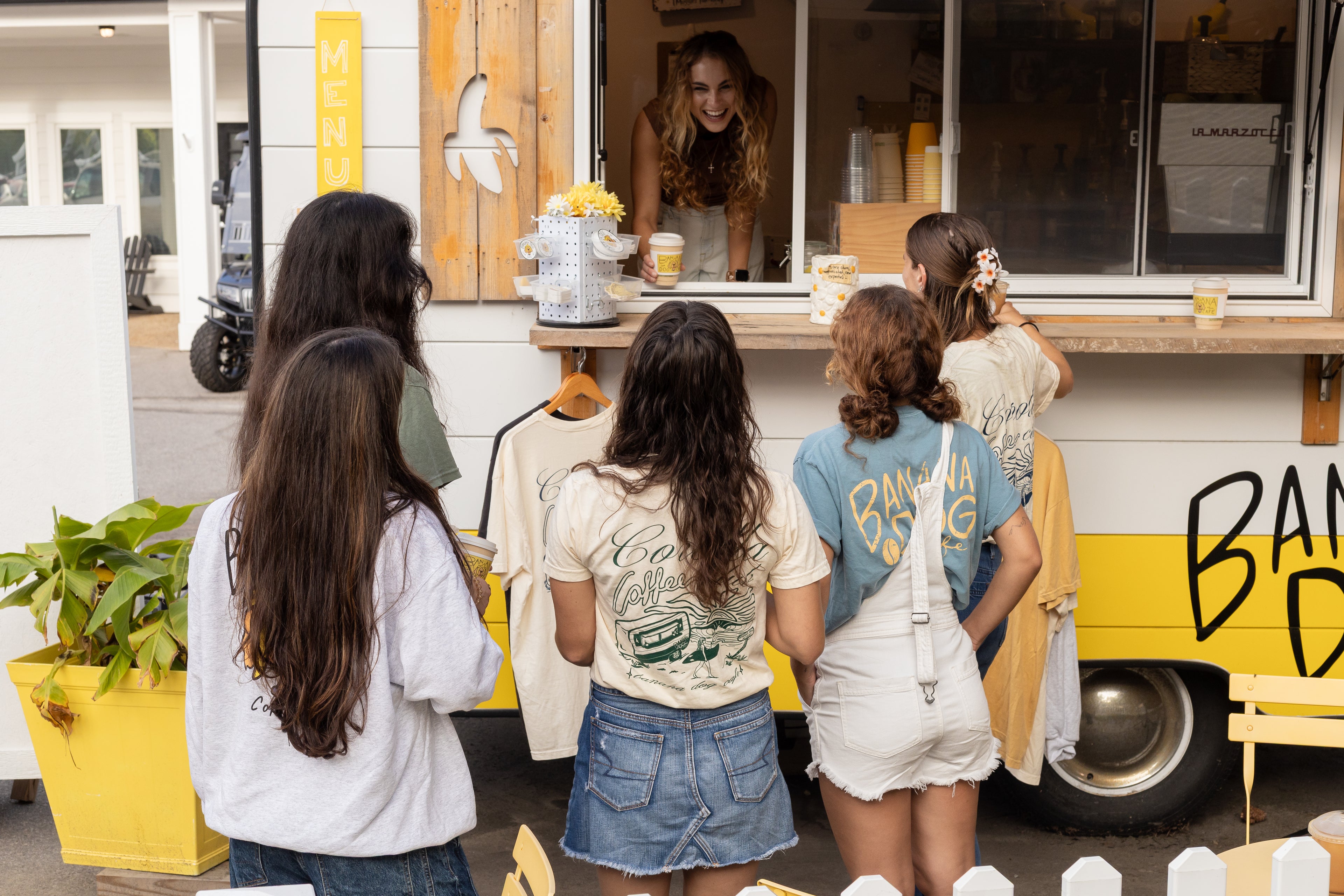 A group of young women stand at the counter of a mobile clothing and food truck, with a woman smiling inside the serving window. The truck is white and yellow with 'BAM' and 'D' visible on the side.
